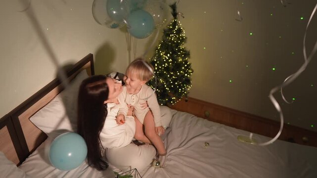A toddler boy cuddles with his mother beneath blue balloons beside the tree in a festive home. The overhead wide view conveys New Year morning, family celebration and family warmth.