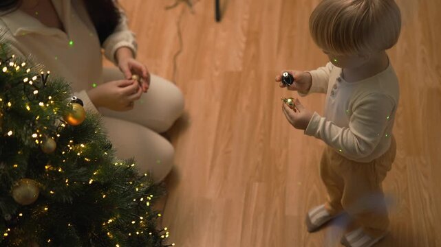 A toddler boy explores shiny ornaments beside a glowing Christmas tree in a festive home. The close candid view conveys childhood curiosity, holiday discovery and family warmth.