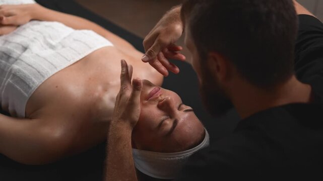 In a wellness session, a massage therapist positions both hands around a woman's head for a calming scalp treatment. The overhead close view highlights stress relief, head care and client care.