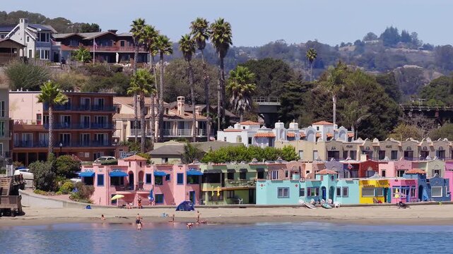 Venetian village beachfront cottages line the sandy shore at Capitola Beach in California on a sunny summer day, with coastal waters, palm trees, and railway trestle bridge visible in background.