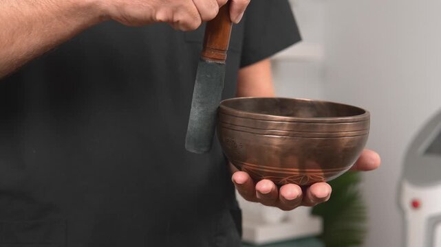 A client relaxes as a wellness practitioner holds a singing bowl and wooden striker during a wellness ritual. This treatment shows sound meditation, mindfulness and wellness practice.