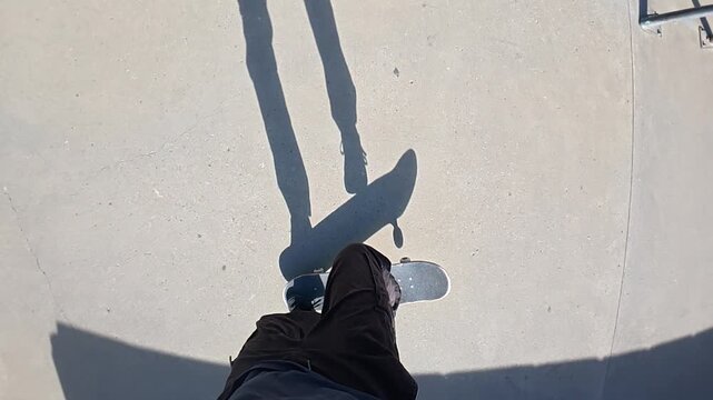 A skateboarder is doing a trick from a POV and slow motion in a skatepark on a sunny day