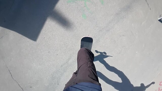 A skateboarder is doing a trick from a POV and slow motion in a skatepark on a sunny day