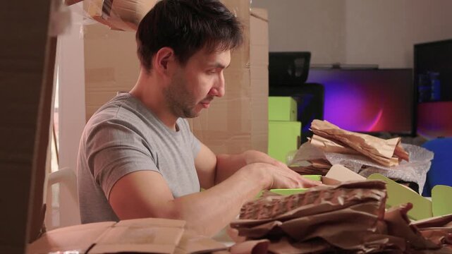 White man wrapping stacked books for shipment at packing table, smoothing kraft paper and placing bundle into green box with bubble wrap and calm, focused expression