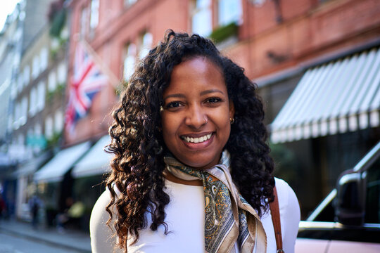 Cheerful millennial woman holds her backpack straps looking at camera on a lively London street, with Union Jack and striped awnings behind her, capturing a bright travel moment with confident energy
