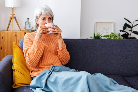 Smiling senior woman relaxing on the sofa, wrapped in a warm blanket and enjoying a hot cup of coffee on winter season