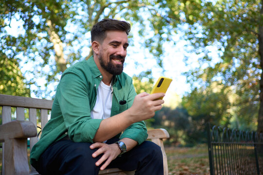 Smiling millennial sits on a park bench in autumn, holding a bright yellow smartphone while checking messages. Urban trees and fallen leaves frame a relaxed, modern lifestyle moment.