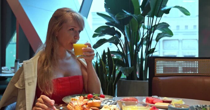 Caucasian woman drinking orange juice during breakfast in modern restaurant. Table with pastries and fruit, green plant background, morning light, lifestyle dining scene.