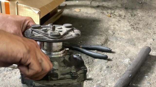 technician is assembling pieces of a broken car air conditioning compressor on the pavement.