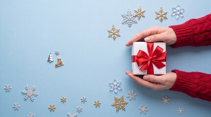 Obraz premium Top View of Woman Hands in Red Knitted Sweater Holding a White Christmas Gift Box with Red Ribbon Bow Surrounded by Snowflake Decorations and Winter Charms on Pastel Blue Background with Copy Space