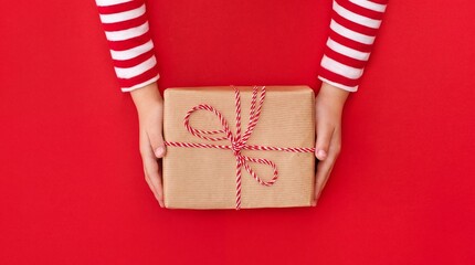 Obraz premium Top view of a child's hands wearing festive red and white striped sleeves holding a handmade gift box wrapped in brown kraft paper and tied with red twine over a vibrant red background layout.