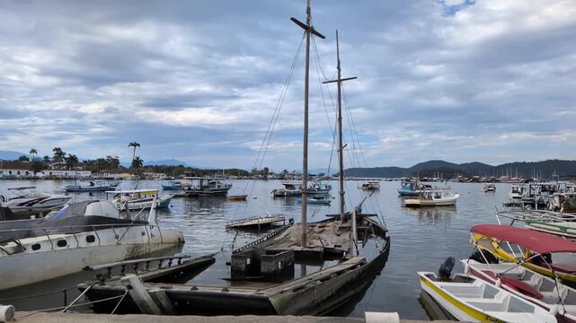 Static slow motion shot of an old sunken wooden boat in the foreground at the Fishermen Port in Paraty, Rio de Janeiro, with other vessels floating under a cloudy sky.