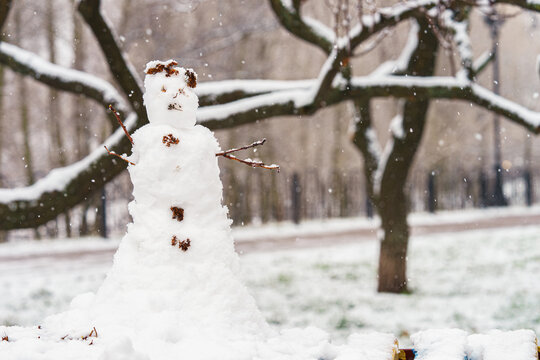 Snowman made of fresh snow standing in a winter park during snowfall, surrounded by snow covered trees and branches, creating a peaceful festive outdoor winter scene.
