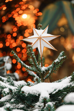 Christmas tree decorated with white star, ornaments and warm fairy lights, displayed outdoors with festive illuminated, light bokeh background and snowy branches. Holiday atmosphere, aesthetic mood