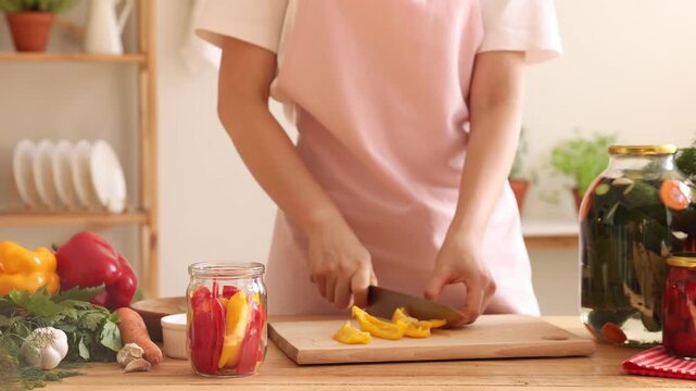 Woman cutting fresh bell pepper and putting it into pickling jar at wooden table indoors, closeup