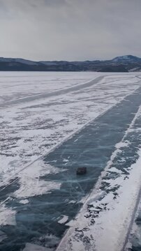 Drone footage of a vehicle driving along a winter ice road on frozen Lake Baikal. Blue transparent ice with cracks and snowfields, mountains on horizon, Russia