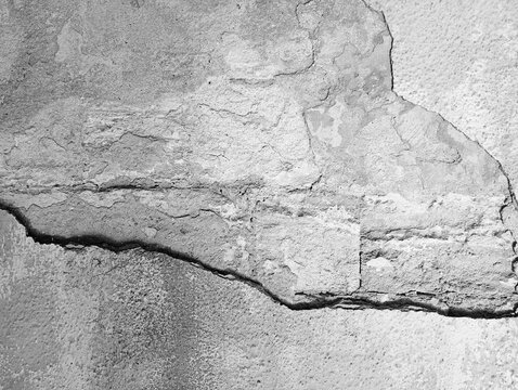 A detailed black and white texture of an old weathered building facade, showing thick concrete plaster layers cracking and breaking away from a rough stone wall, 