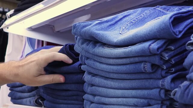 Man choosing blue denim jeans from folded stacks on display in clothing store. Close-up shopping for casual fashion apparel in retail.