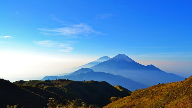 Layered mountain silhouettes with volcano peaks under clear blue sky, golden grassy hills in foreground, serene morning landscape.
