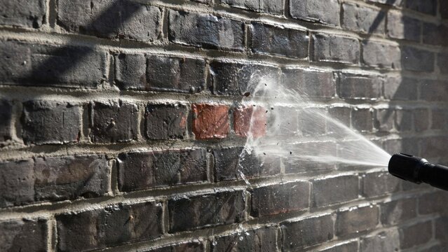 Pressure water spraying on dark brick wall revealing original red bricks from under layers of paint during cleaning.