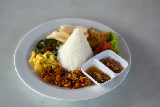 Indonesian cone shaped rice tumpeng with side dishes on white plate.
