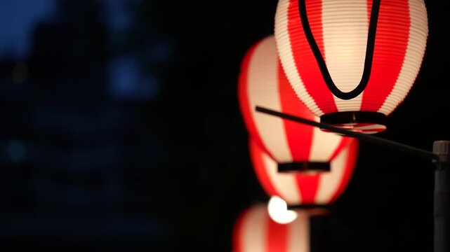 cinematic close-up shot of illuminated traditional japanese red and white paper lanterns hanging in a row at night with a dark blurred background