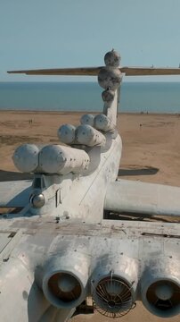 Aerial view of an abandoned Soviet-era ekranoplan ground effect aircraft resting on a sandy shoreline, showing large wings and multiple jet engines in daylight