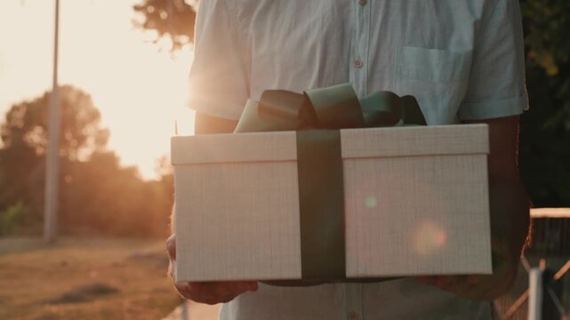 A man in a white shirt holds a beautifully wrapped gift box with a green ribbon, walking towards the camera with the sun setting in the background, creating a warm and inviting atmosphere