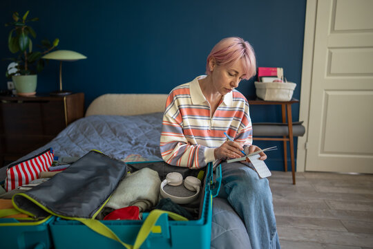 Focused woman writing packing checklist in notepad while preparing suitcase for solo travel, thoughtful female sitting on bed in bedroom organizing luggage for vacation trip marking in to-do list.