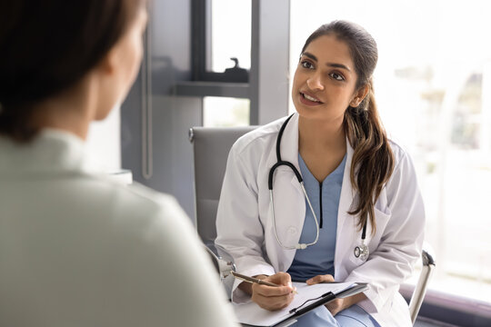 Indian woman doctor in uniform asking female patient about disease symptoms or health concerns seated in front of her, holding clipboard and pen, writing notes during medical consultation at hospital