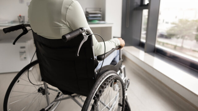 Close up cropped image rearview, unrecognizable young woman with disability sitting in wheelchair looking out window in medical office, getting accessible healthcare services in rehabilitation center