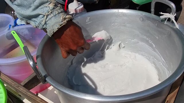 Video of Hand stirs Indonesian sweet coconut rice porridge in large metal pot. Sunlight highlights the creamy texture of the batter. Surrounding containers hold colorful liquids and tools. 4K Cuisine