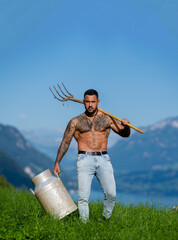 Obraz premium Farmer carrying milk can on grassy hillside. Handsome farmer with pitchfork beside mountain lake. Farmer with bare chest in rural portrait. Tattooed dairy farmer carrying farm tool outdoors.