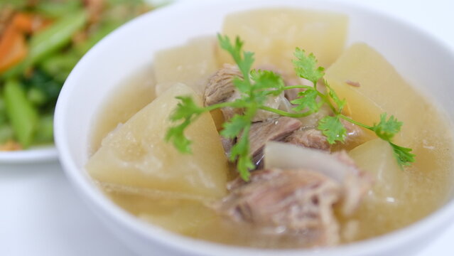  A white bowl of clear winter melon (white gourd) tender pork rib soup with blurred of stir-fried oyster sauce vegetable in the background.