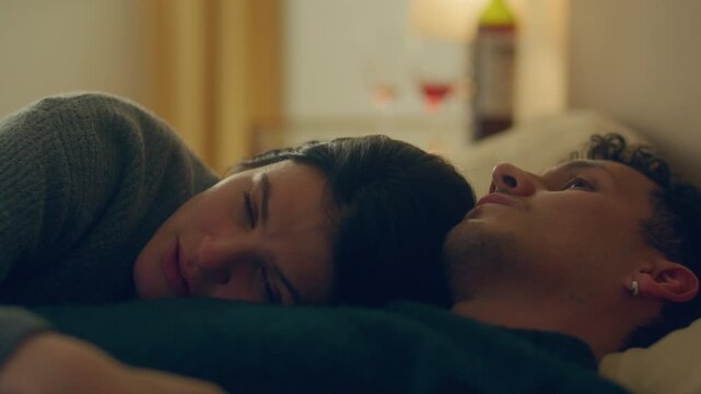 Sad young woman with tearful eyes lying down in bed, resting her head on partners chest and talking vulnerably to him after argument