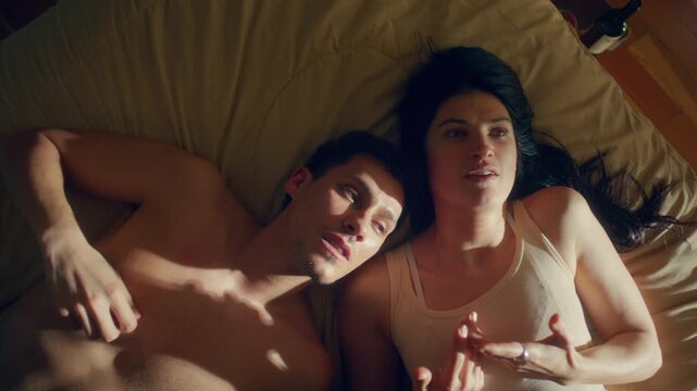 Upset young woman and shirtless man lying side by side in bed, speaking emotionally during heated argument. High angle view