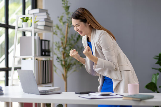 Asian businesswoman raising her arms in excitement while achieving a business target using her laptop in the office, expressing joy and accomplishment.