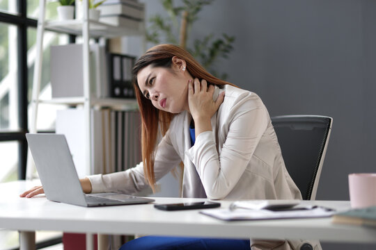 Stressed businesswoman massaging her neck, experiencing discomfort and stiffness after long hours of working on a laptop in office.
