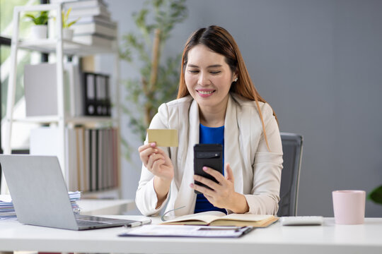Asian businesswoman smiling and holding credit card while using smartphone for online shopping and entering information, laptop and notebook on desk in office.