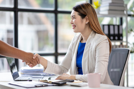 Asian businesswoman sitting at her desk shaking hands with a client after closing a deal in office, concept of successful negotiation and agreement.