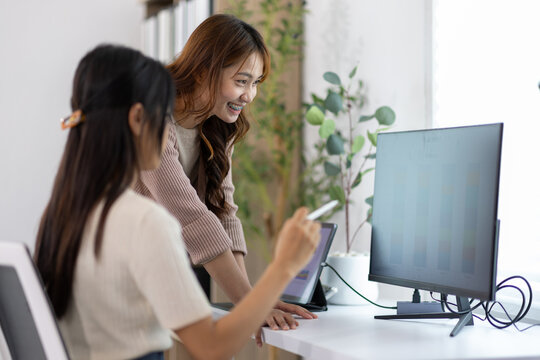 Two young businesswomen collaborating on a project, pointing at a computer screen while discussing innovative ideas in office, teamwork and communication.