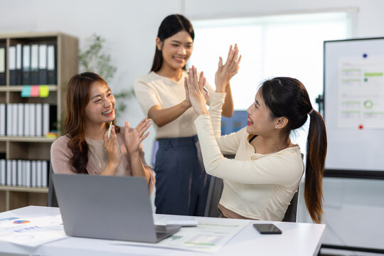Three Asian businesswomen are giving high five to each other, celebrating the success of project and looking happy and satisfied in modern office.