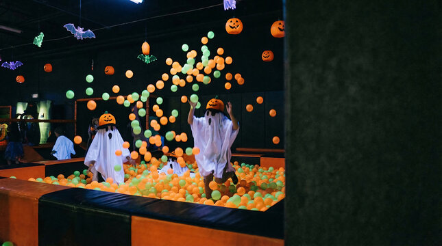 Excited children in ghost and pumpkin costumes celebrating a festive holiday in an indoor play area, surrounded by colorful balls and decorations.