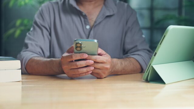 Mid adult man holding mobile phone sitting at wooden desk near tablet computer stack books indoors.