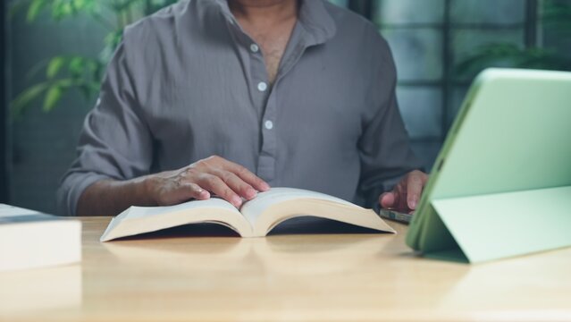 Adult man reads printed textbook beside digital tablet at wooden desk indoors, conducting research, studying course literature, preparing academic assignment carefully.