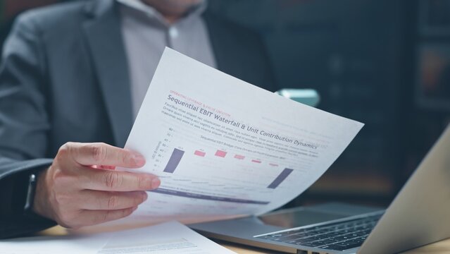Businessman reviewing financial document showing sequential ebit waterfall chart beside laptop computer in corporate office setting.