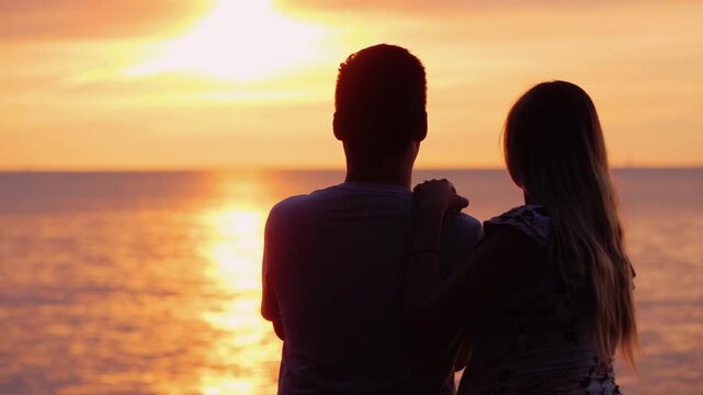 Silhouette of romantic young couple watching orange sunset together over ocean waves and horizon.