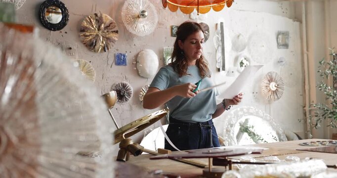 Female artisan measuring and cutting paper templates for decorative glass elements of bespoke chandelier constructing layout sample and preparing technical design before fabrication in craft workshop