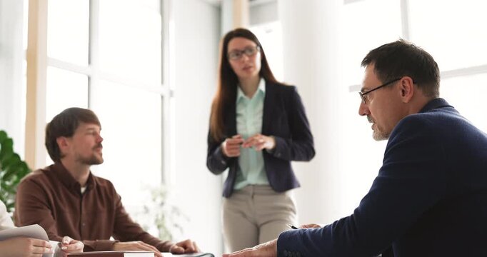 Experienced middle aged male executive speaking during corporate team discussion offering expertise to female leader and coworker group sharing professional knowledge with younger staff at office desk