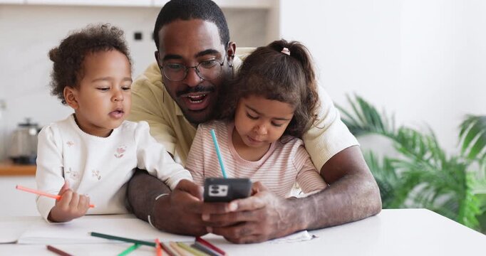 African father guiding two little daughters with mobile art tutorial on smartphone helping children draw pictures with colored pencils and follow step by step online sketching lesson at kitchen table
